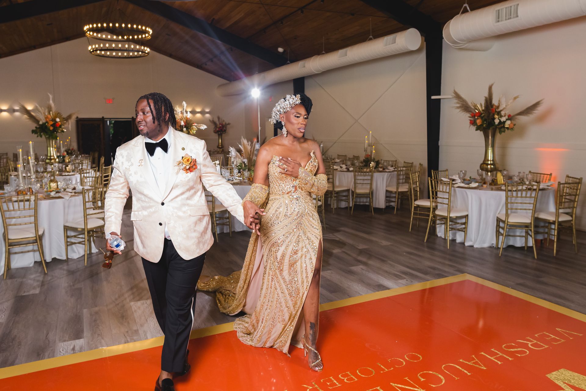 Couple walking hand in hand at a decorated indoor reception, with tables and floral arrangements in the background