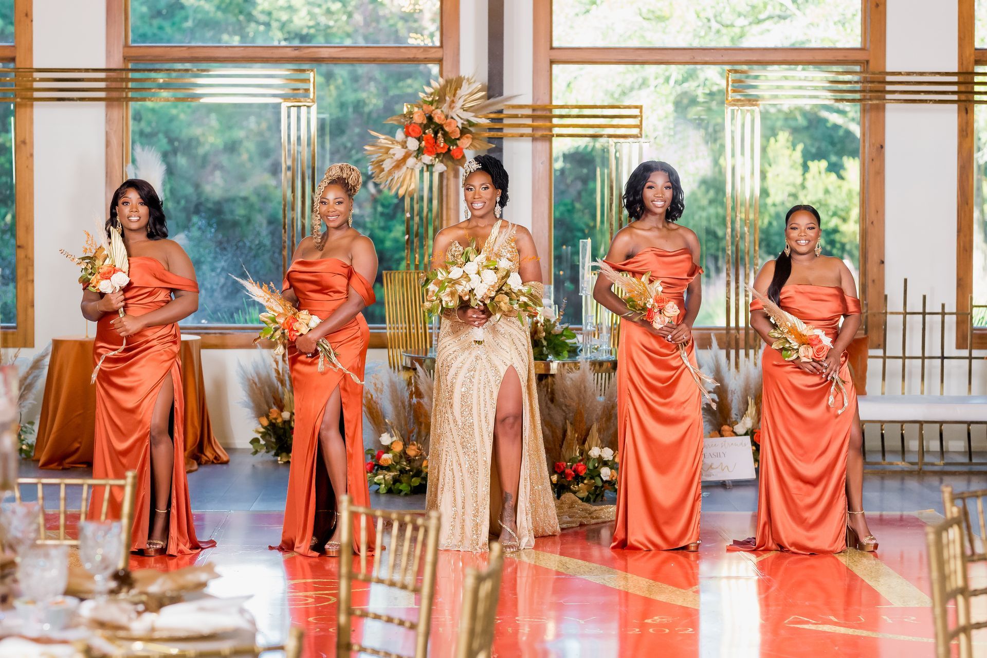 Six women in orange dresses stand with bouquets in a bright, elegant hall.