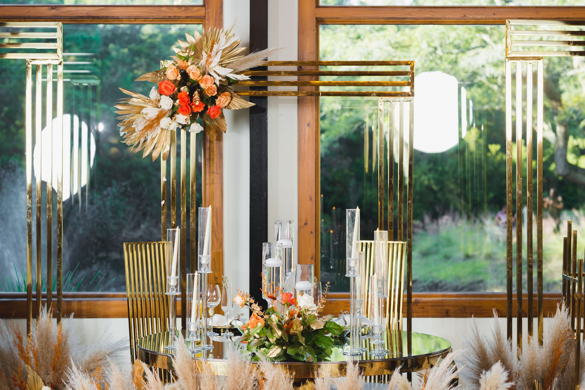 Elegant table setting with tall gold candles and orange-white floral arrangements by a large window