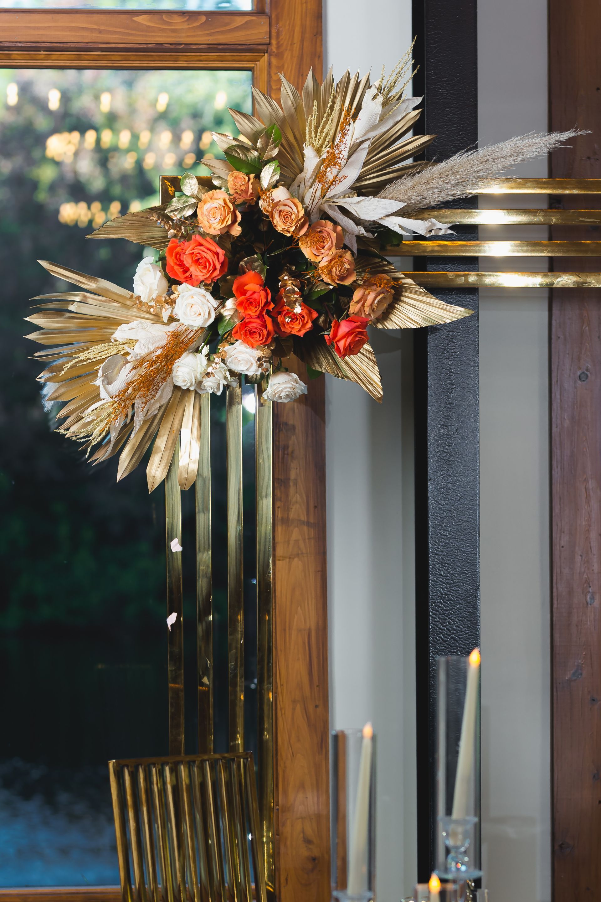 Floral arrangement with white, orange, and red flowers in a brass vase by a window or mirror.