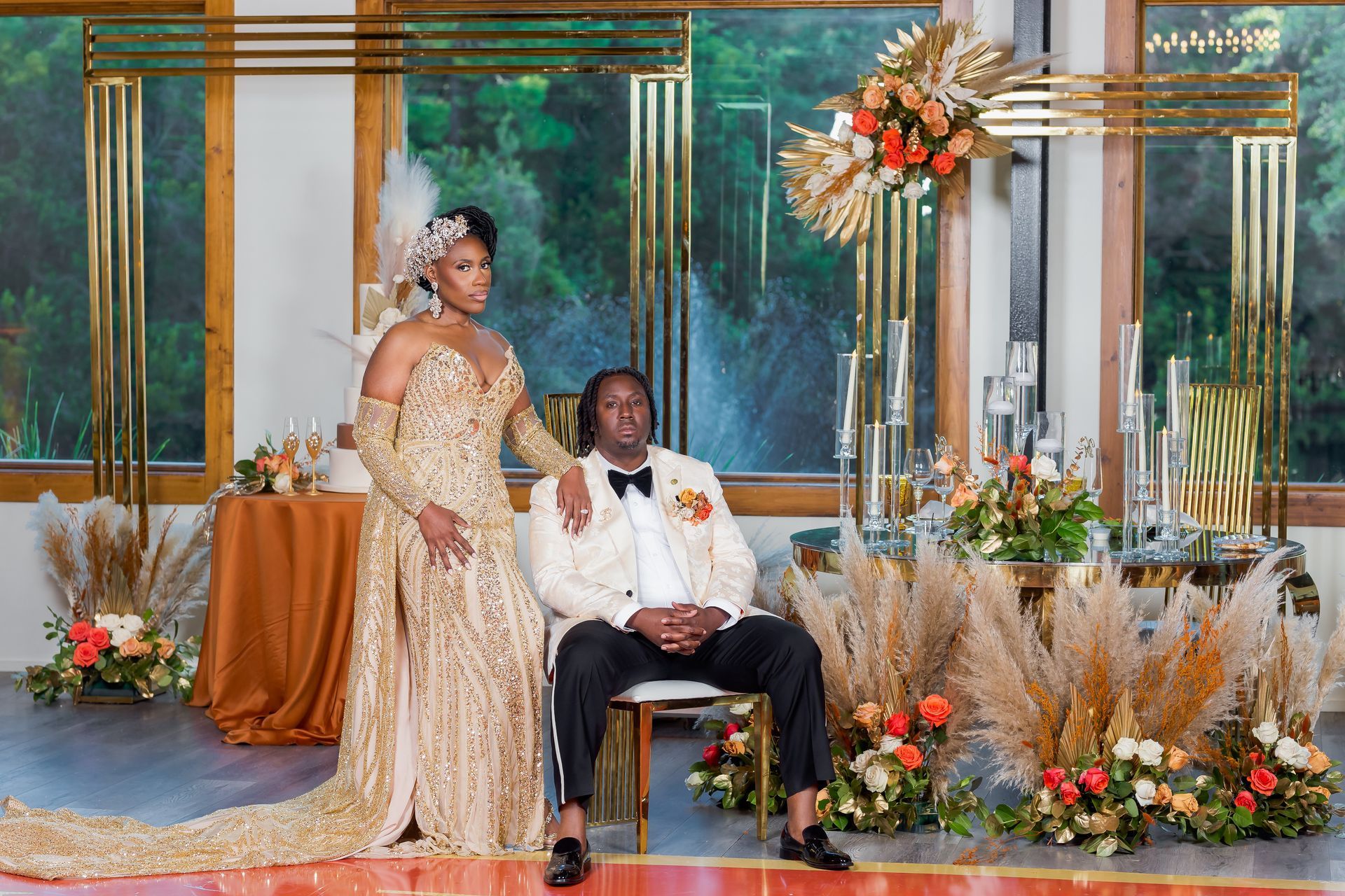 Couple in gold formalwear at an elegant floral wedding stage, with the man seated and the woman standing beside him
