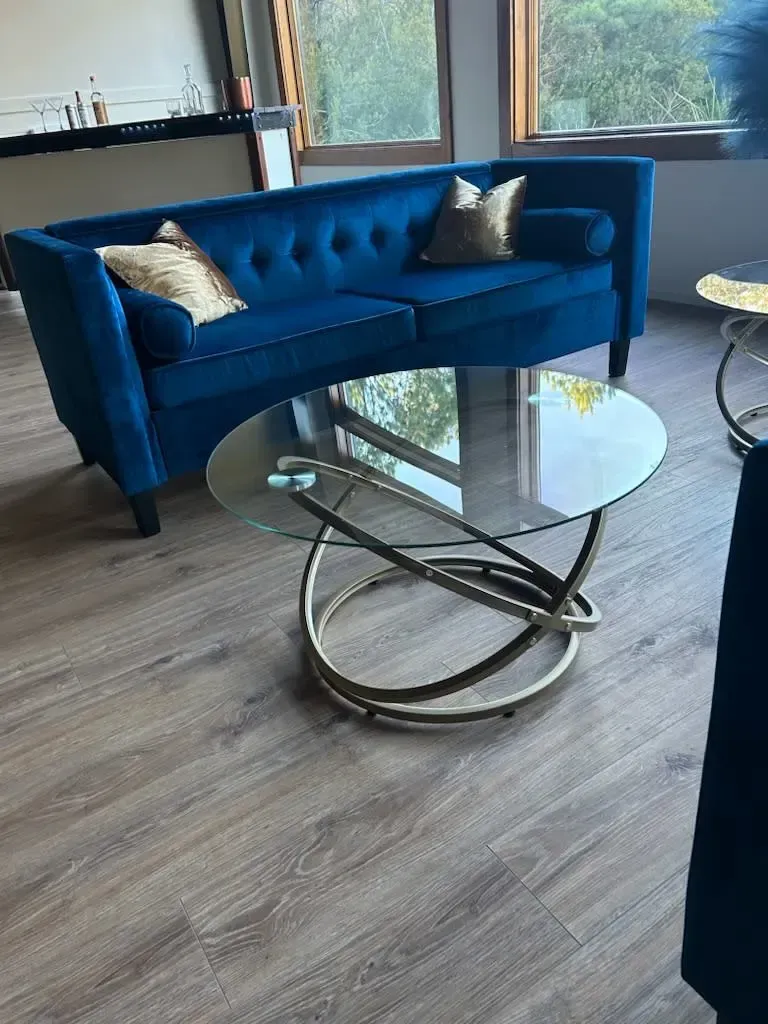 Blue loveseat and glass coffee table in a modern room with wood flooring and large windows.
