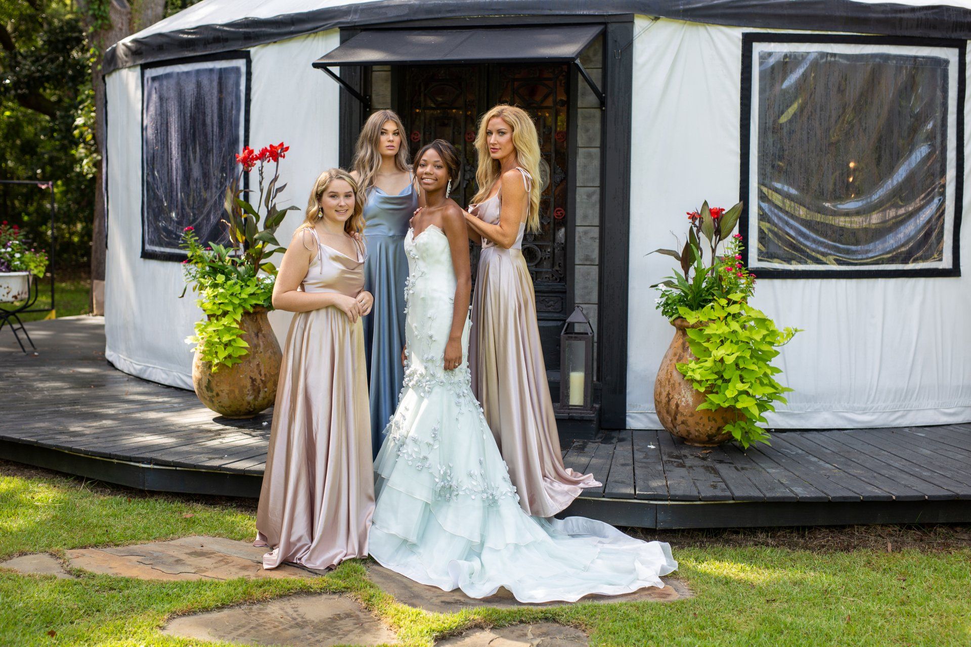 Four women in formal dresses posing outside a white building with flowers by the entrance.