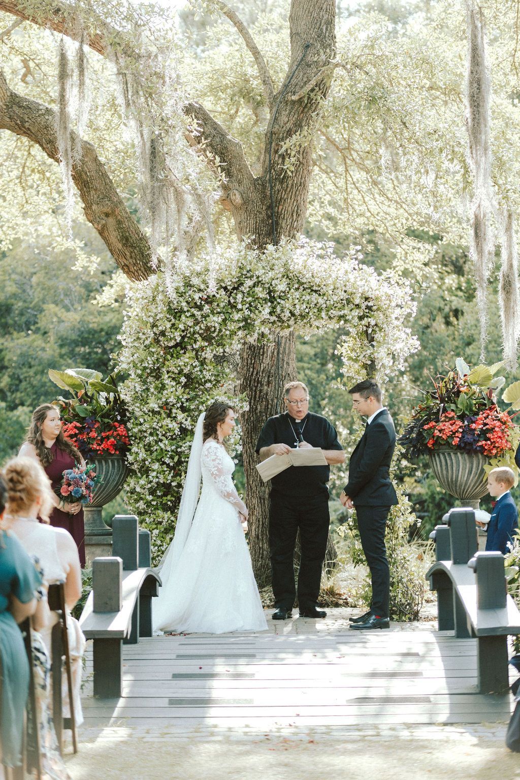 Outdoor wedding ceremony under trees, bride and groom at altar with officiant and floral arch