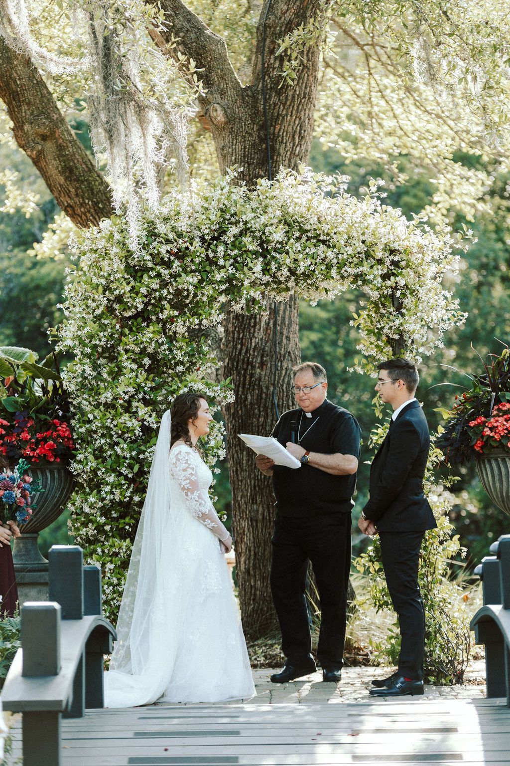 Outdoor wedding ceremony beneath a flower arch, with bride, groom, and officiant standing among trees.