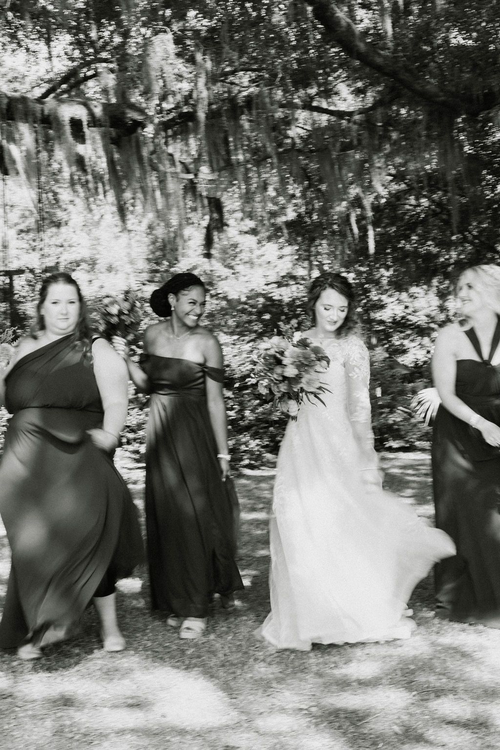 Four people in formal dresses standing outdoors in front of a rock wall and trees