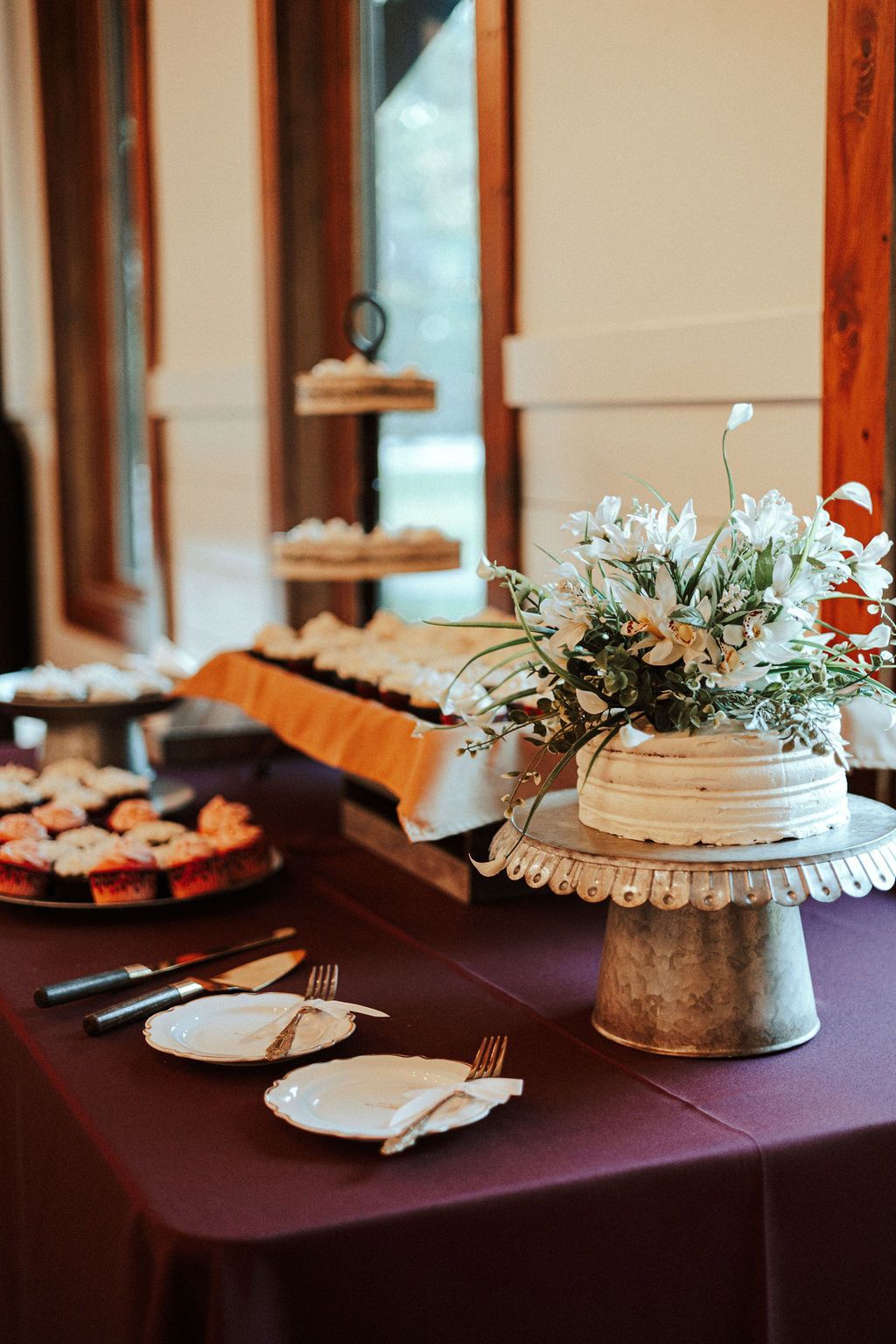 Floral table display with a wedding cake, desserts, and white napkins on a burgundy tablecloth