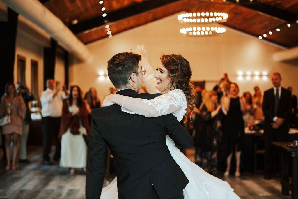 Couple dancing at a wedding reception, surrounded by applauding guests in a warmly lit hall