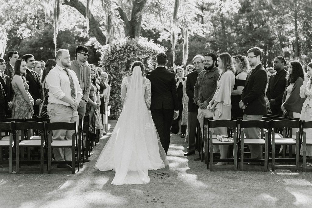 Bride in a long veil walks down an outdoor aisle between seated wedding guests under trees.