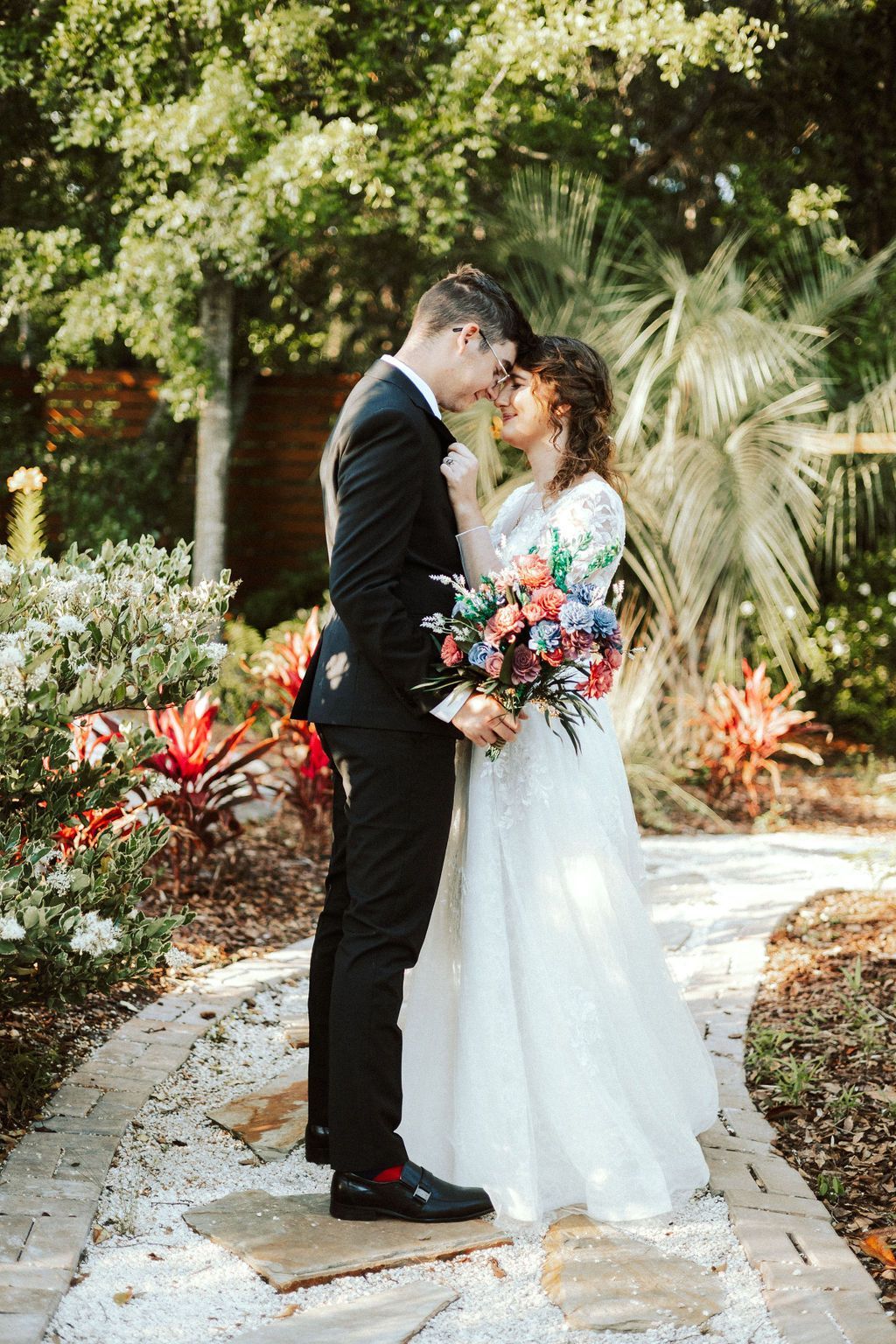 Couple in wedding attire kissing on a garden path surrounded by flowers and greenery