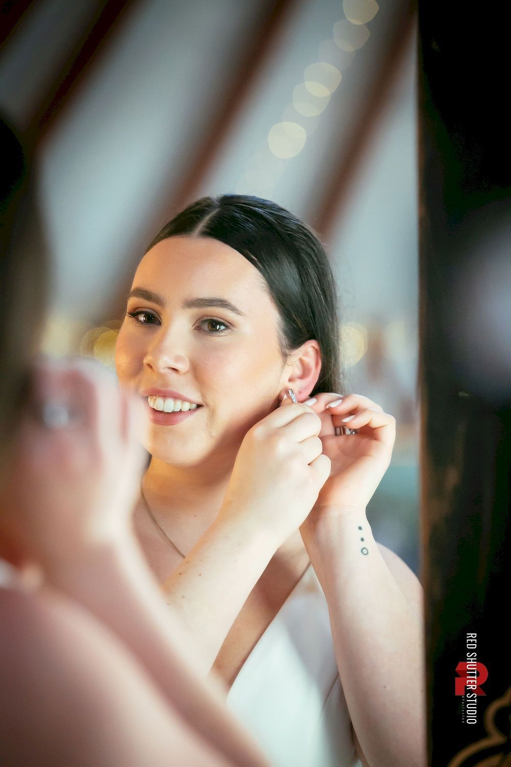 Woman fastening an earring while smiling in a mirror, with soft warm light and blurred background.