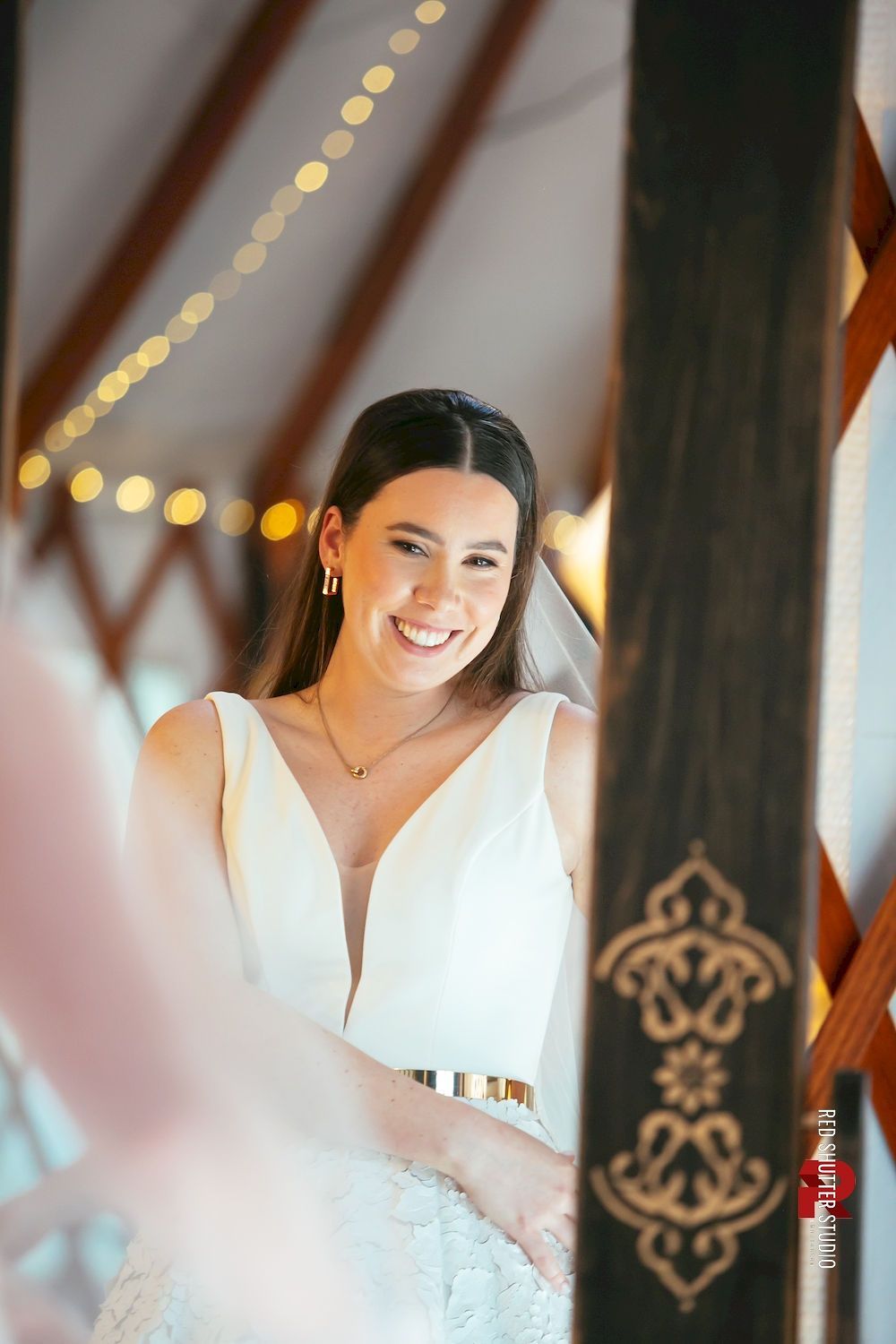 Smiling bride in a white dress standing by a window, framed by warm string lights