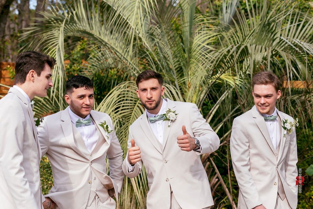 Four men in white suits posing outdoors at a wedding, with one giving a thumbs-up.