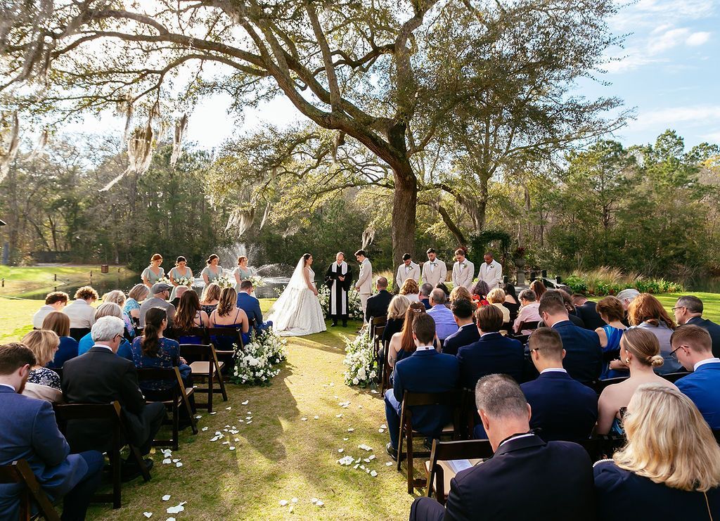 Outdoor wedding ceremony under a large tree with guests seated along a grassy aisle