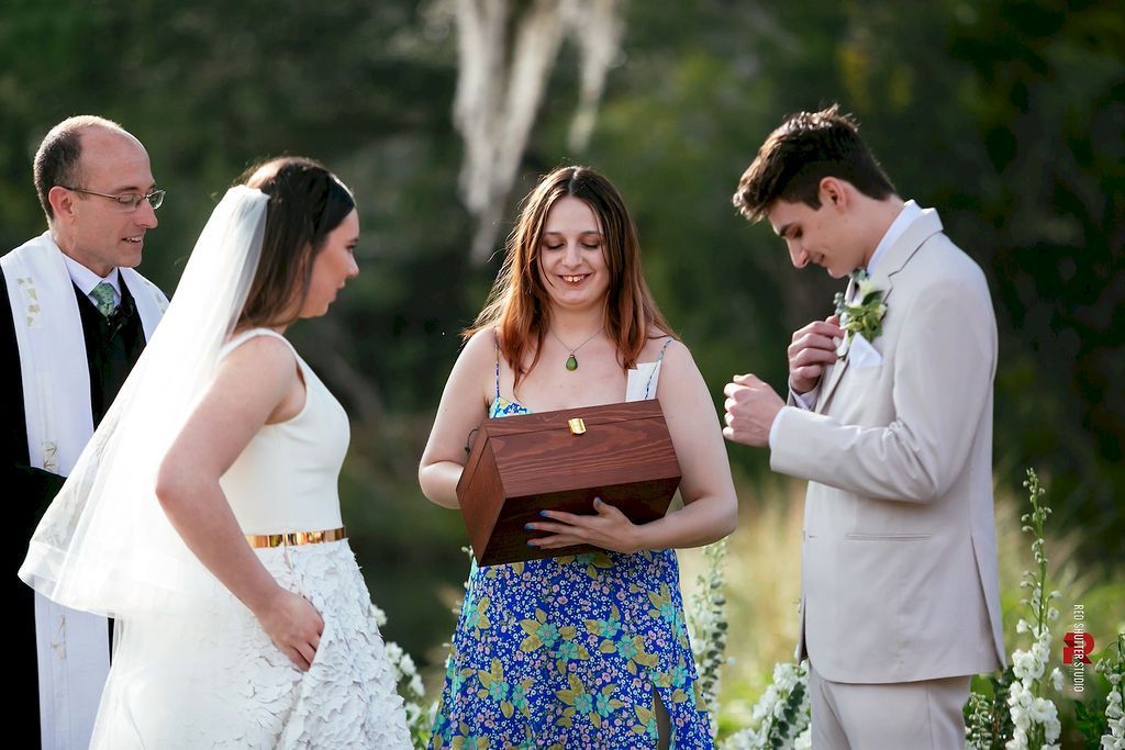 Outdoor wedding ceremony with officiant holding a book, bride and groom facing each other, and a guest nearby