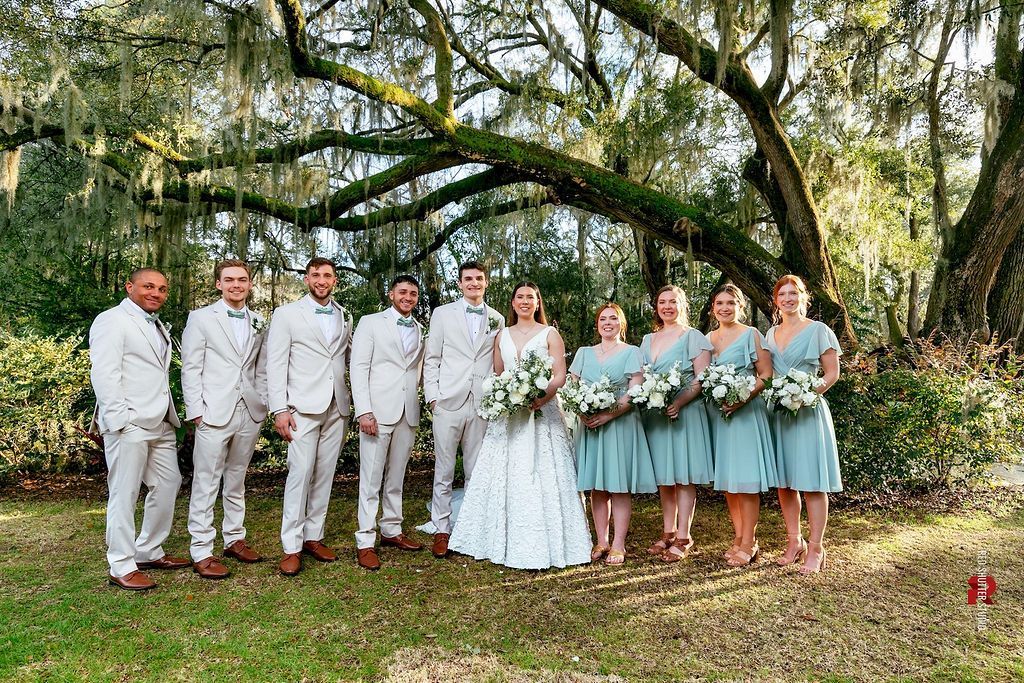 Wedding party posing outdoors under a large oak tree; bride in white, attendants in light blue and gray suits.