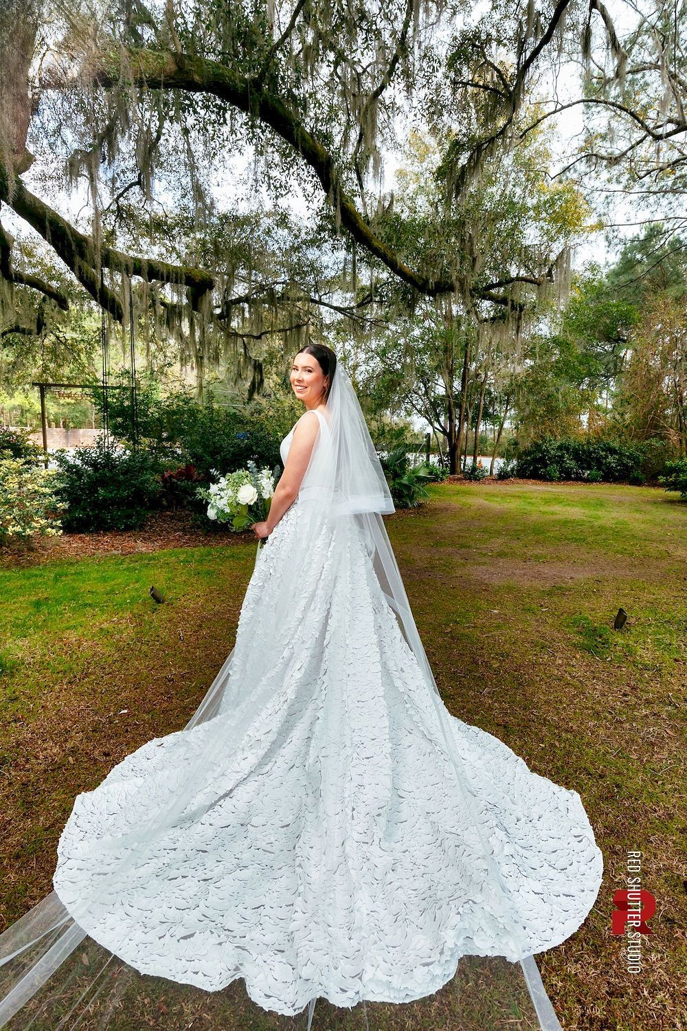 Bride in a long white gown with a flowing train, standing in a leafy garden under trees.