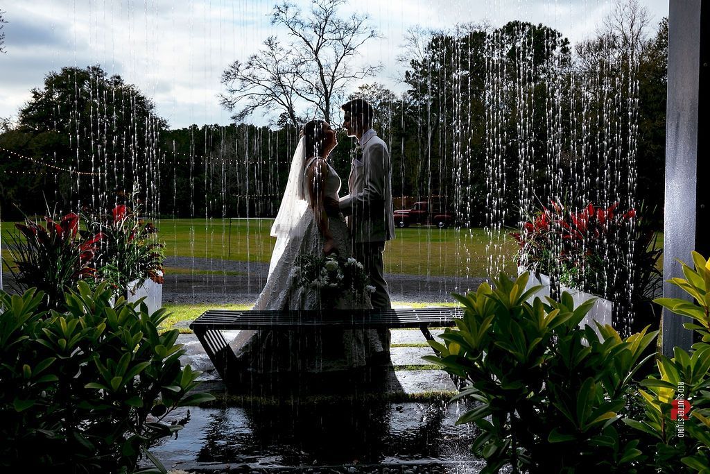 Bride and groom under a fountain in a garden, seen through falling water and framed by flowers and trees
