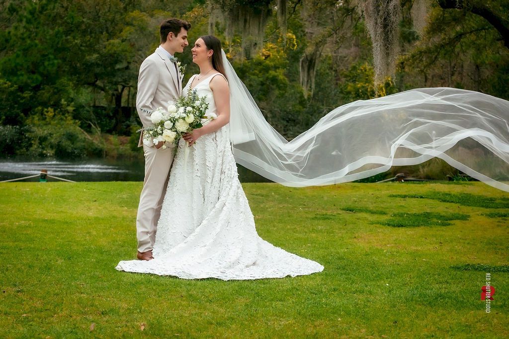 Bride and groom in white wedding attire standing on grass, with the bride’s veil flowing behind them near a river.