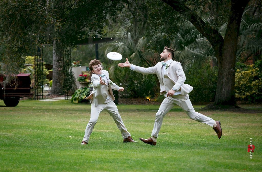 Two people in white fencing outfits spar outdoors, one lunging for a flying disc on a grassy lawn.