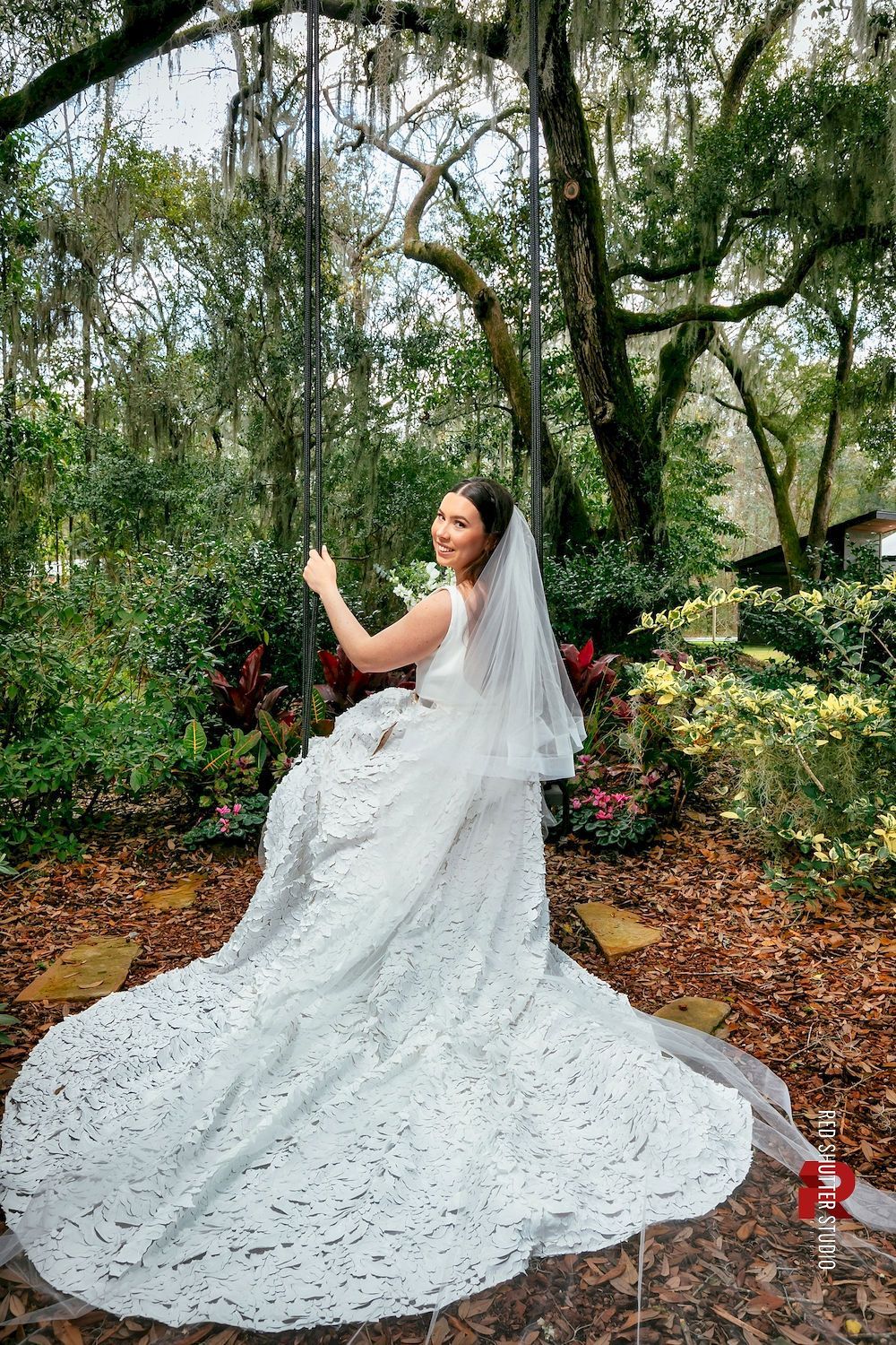 Bride in a white gown and veil standing in a wooded garden, holding up her phone.