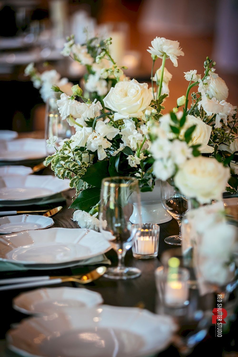Elegant banquet table with white floral centerpieces, glass votives, and white plates set for dining