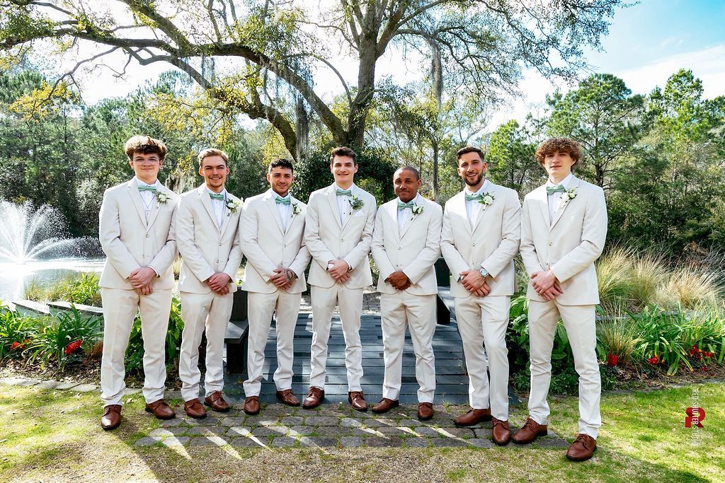 Seven men in cream suits standing together outdoors in a garden, with trees and a fountain behind them