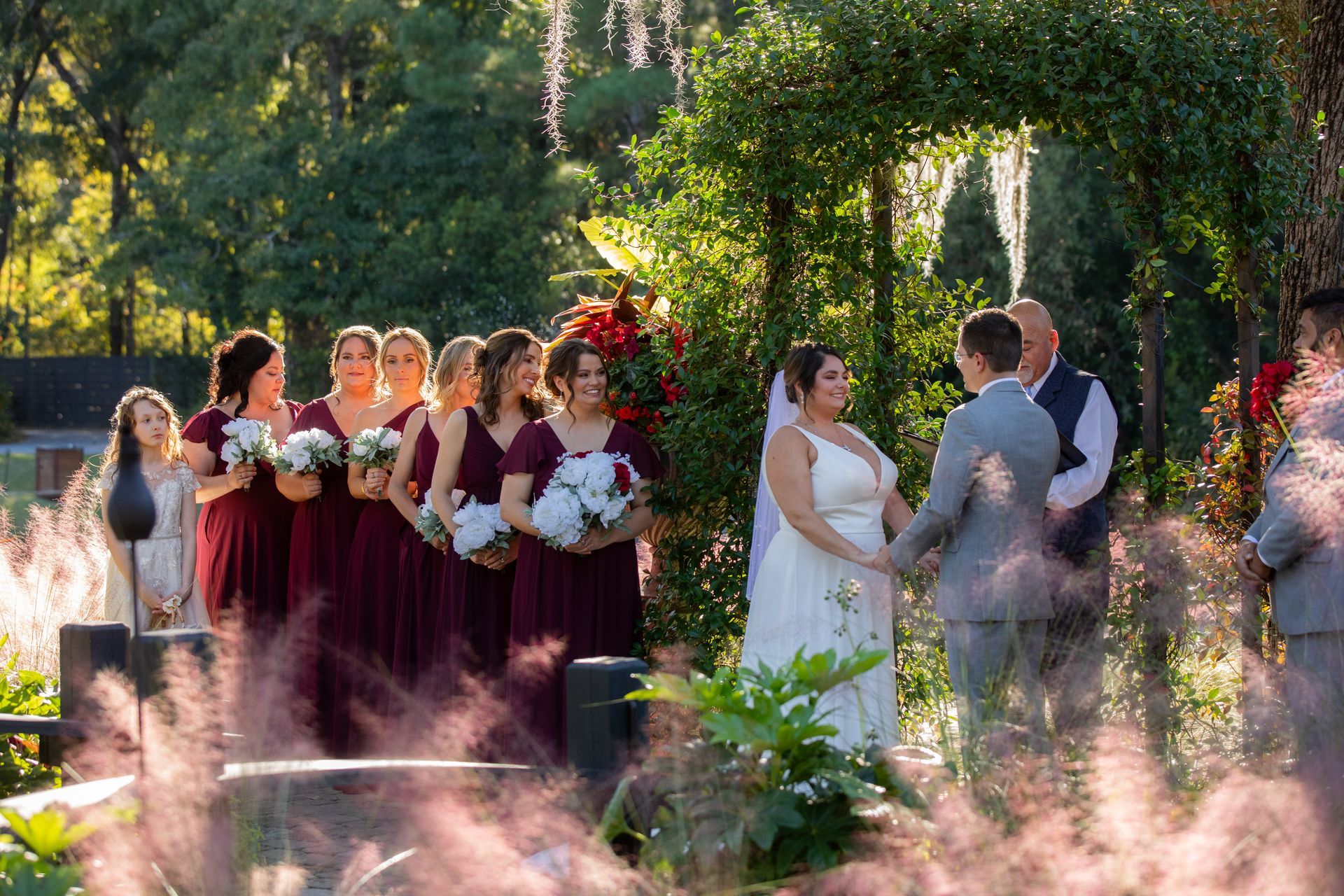 Wedding party outdoors by a flower arch, with bride, groom, and attendants in burgundy dresses and suits