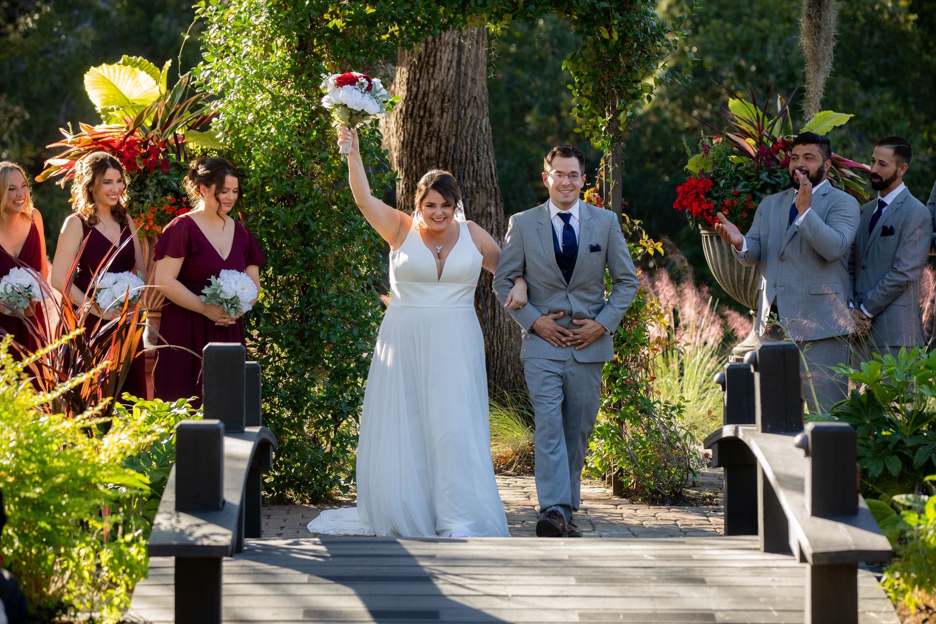 Bride and groom walking down an outdoor aisle, bride raising bouquet amid guests and flowers