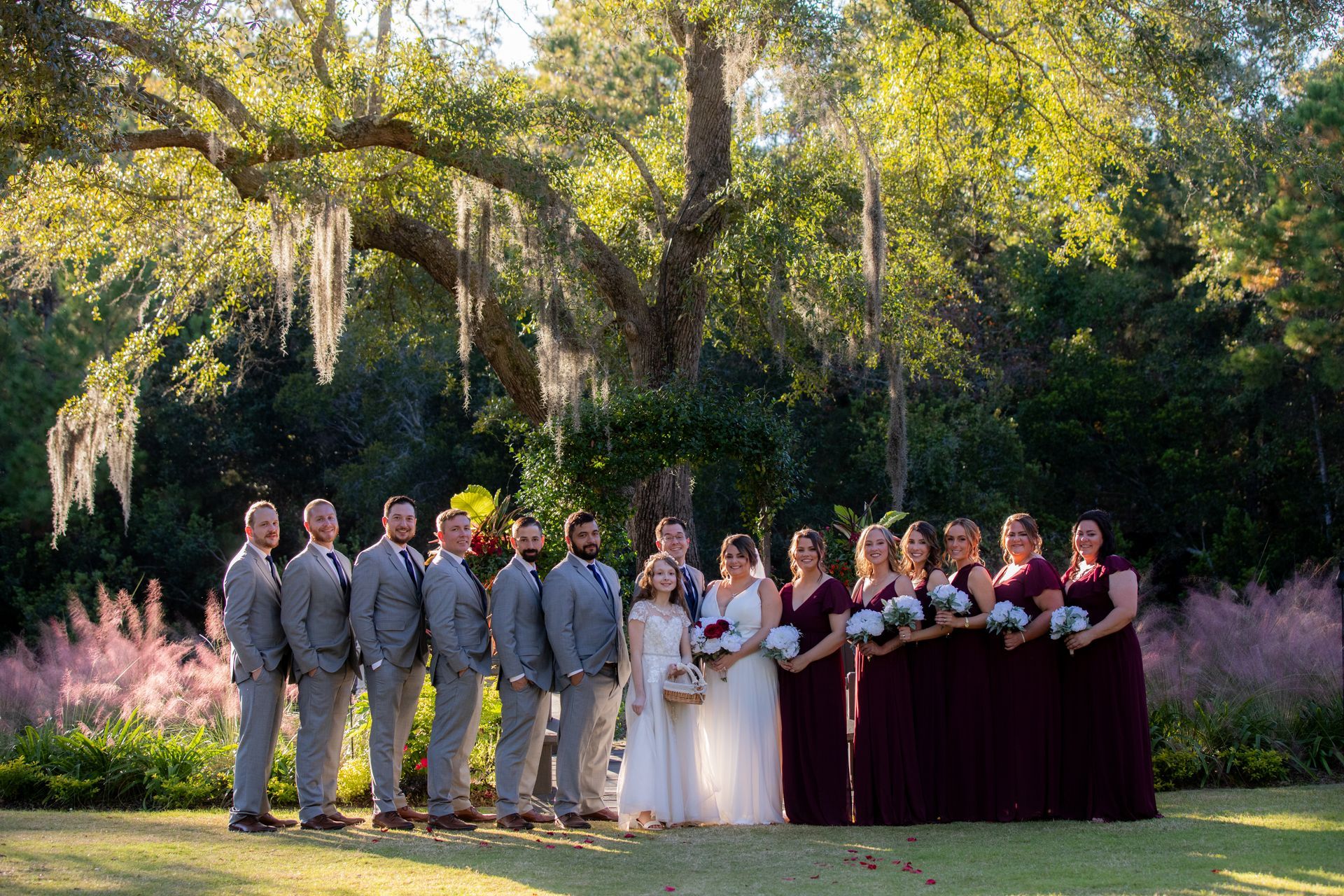 Wedding party posing outdoors under a large tree, bridesmaids in burgundy and groomsmen in gray suits