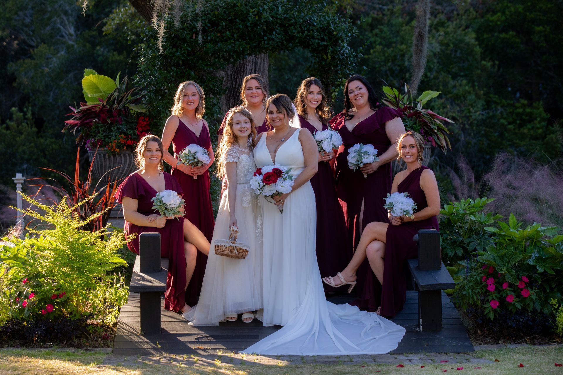 Wedding party posing outdoors in burgundy dresses and white gown, holding bouquets on stone steps.
