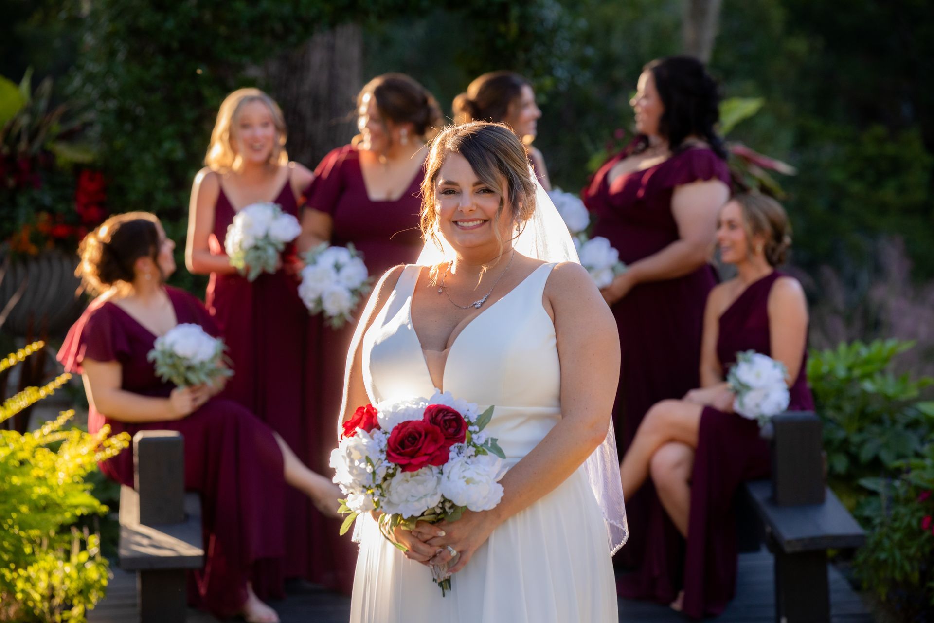 Bride in white dress holding bouquet, with bridesmaids in burgundy dresses seated behind her outdoors.
