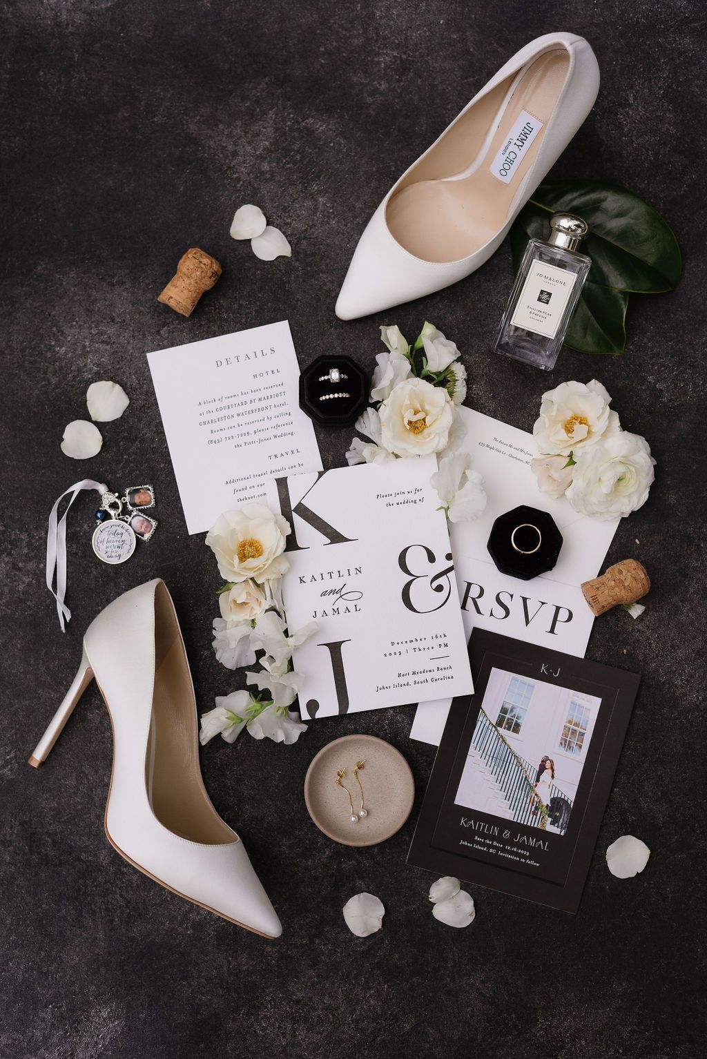 Flat lay of white wedding stationery, heels, flowers, and candle on a dark background