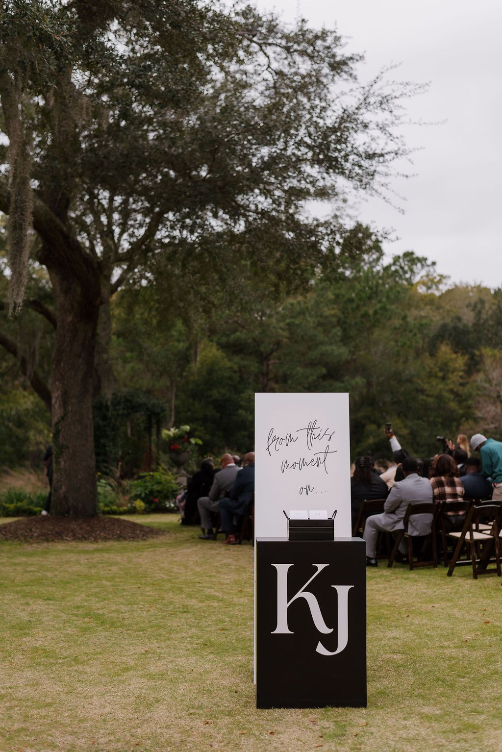 Outdoor wedding setup with guests seated near a white and black “kj” sign on a lawn