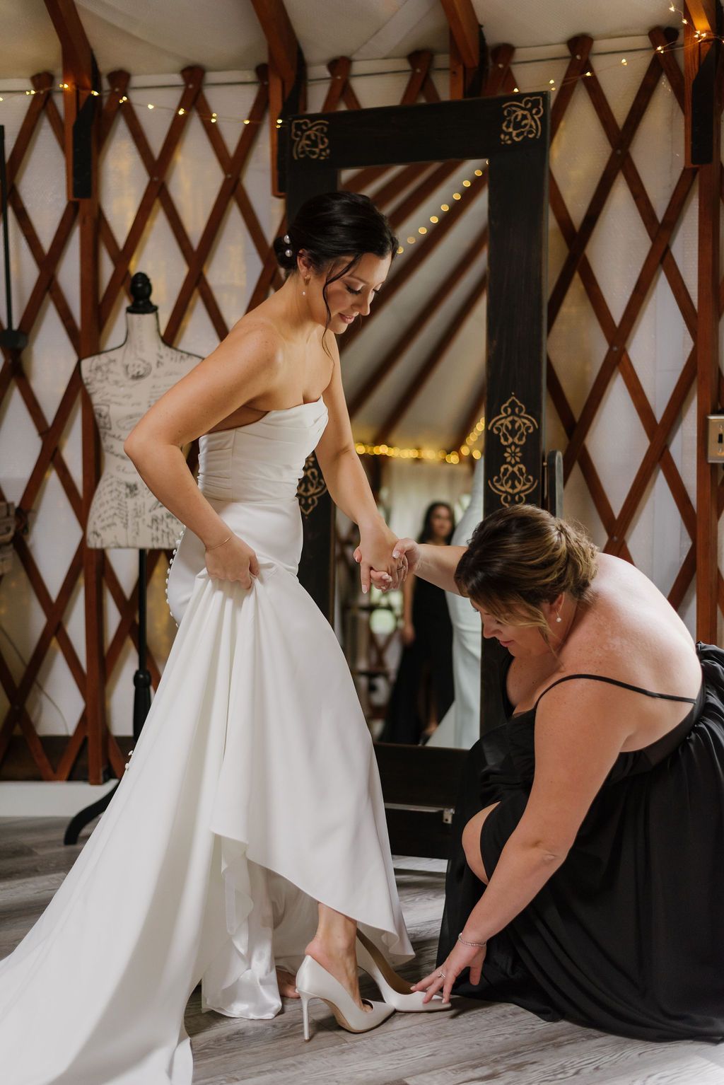 Bride in white gown being helped with her dress by a woman in a black dress indoors