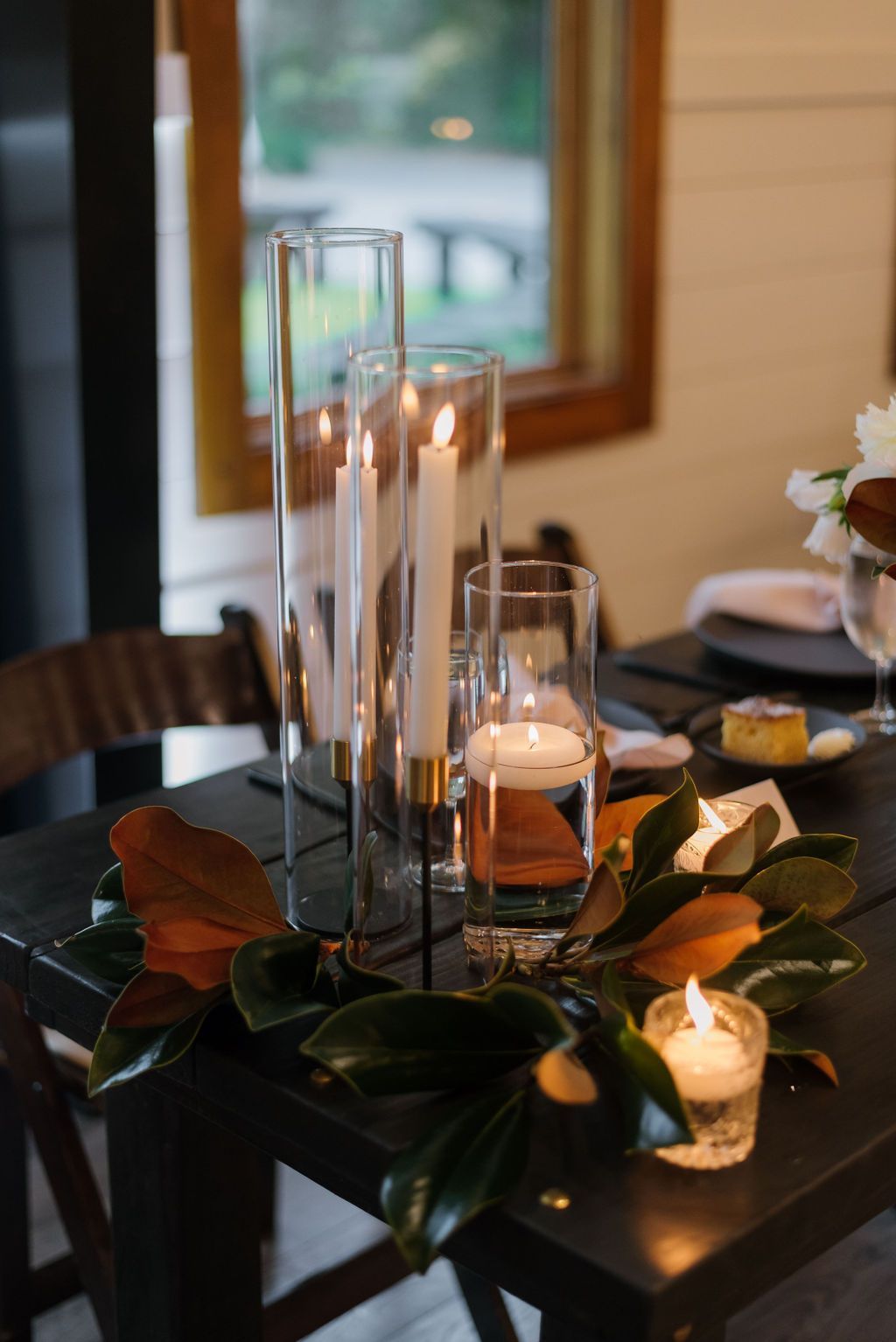 Candlelit dining table centerpiece with glass vases, candles, and autumn leaves beside a window