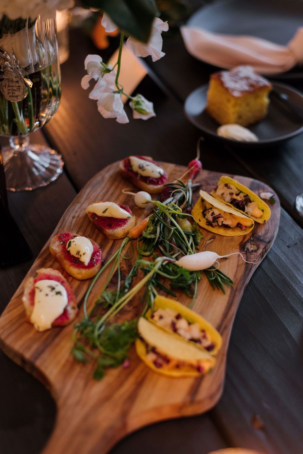 Artisan appetizer platter on a wooden board with sliced fruit, herbs, and edible flowers on a dark table