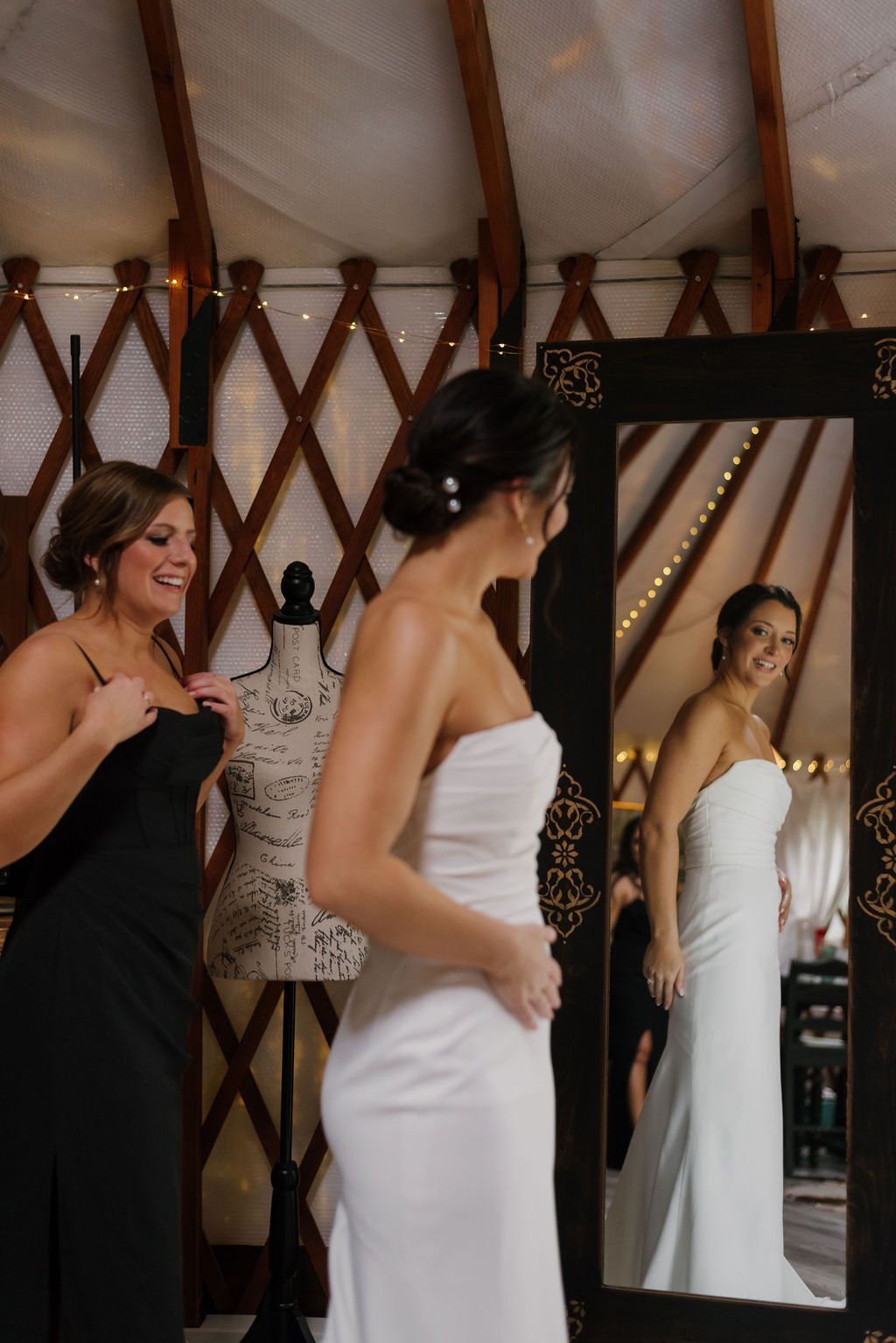 Woman in a white strapless dress stands by a mirror, while another woman adjusts her attire in a rustic room.