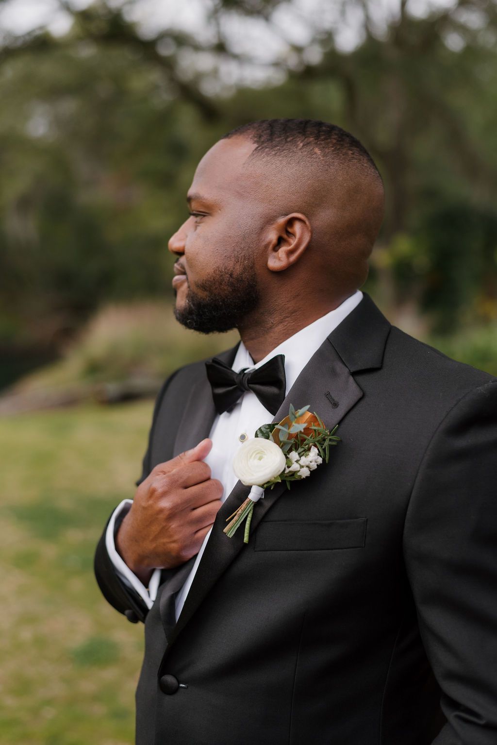 Man in a black tuxedo adjusting his boutonniere outdoors with blurred greenery behind him