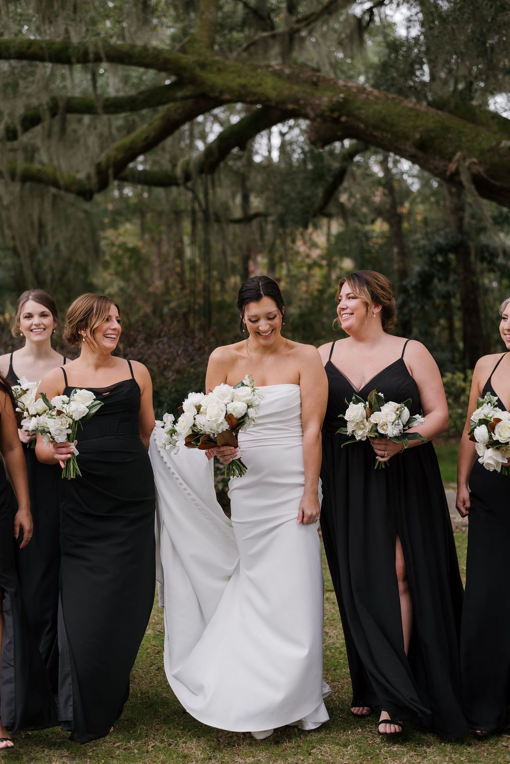 Bride in white dress holding bouquet, flanked by bridesmaids in black dresses outdoors under trees.