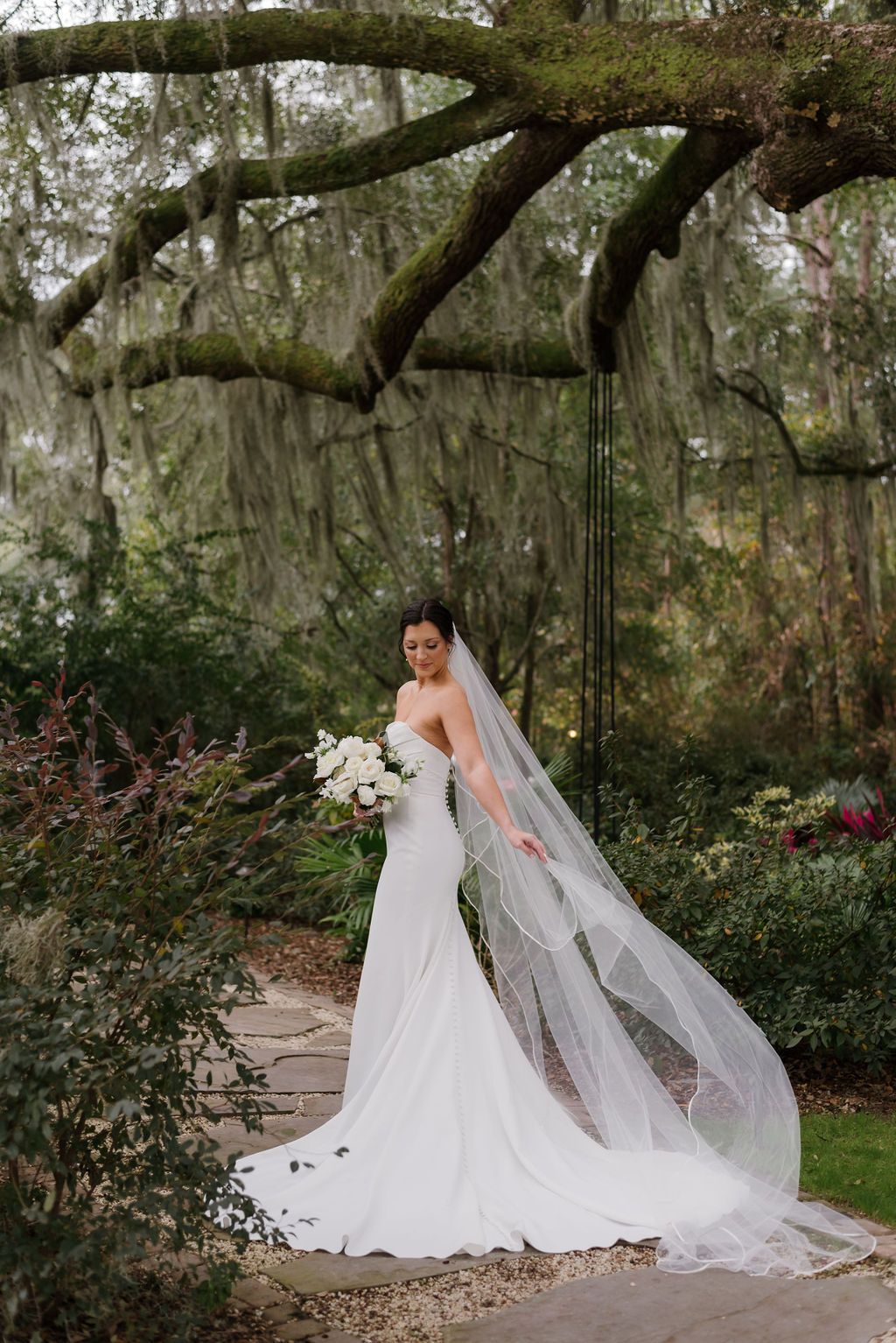 Bride in a white gown with a long veil holding a bouquet in a garden under sprawling tree branches