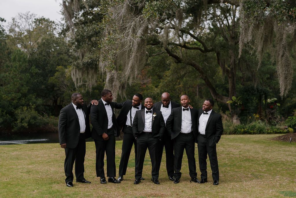 Six men in black suits stand together on a grassy lawn under trees.