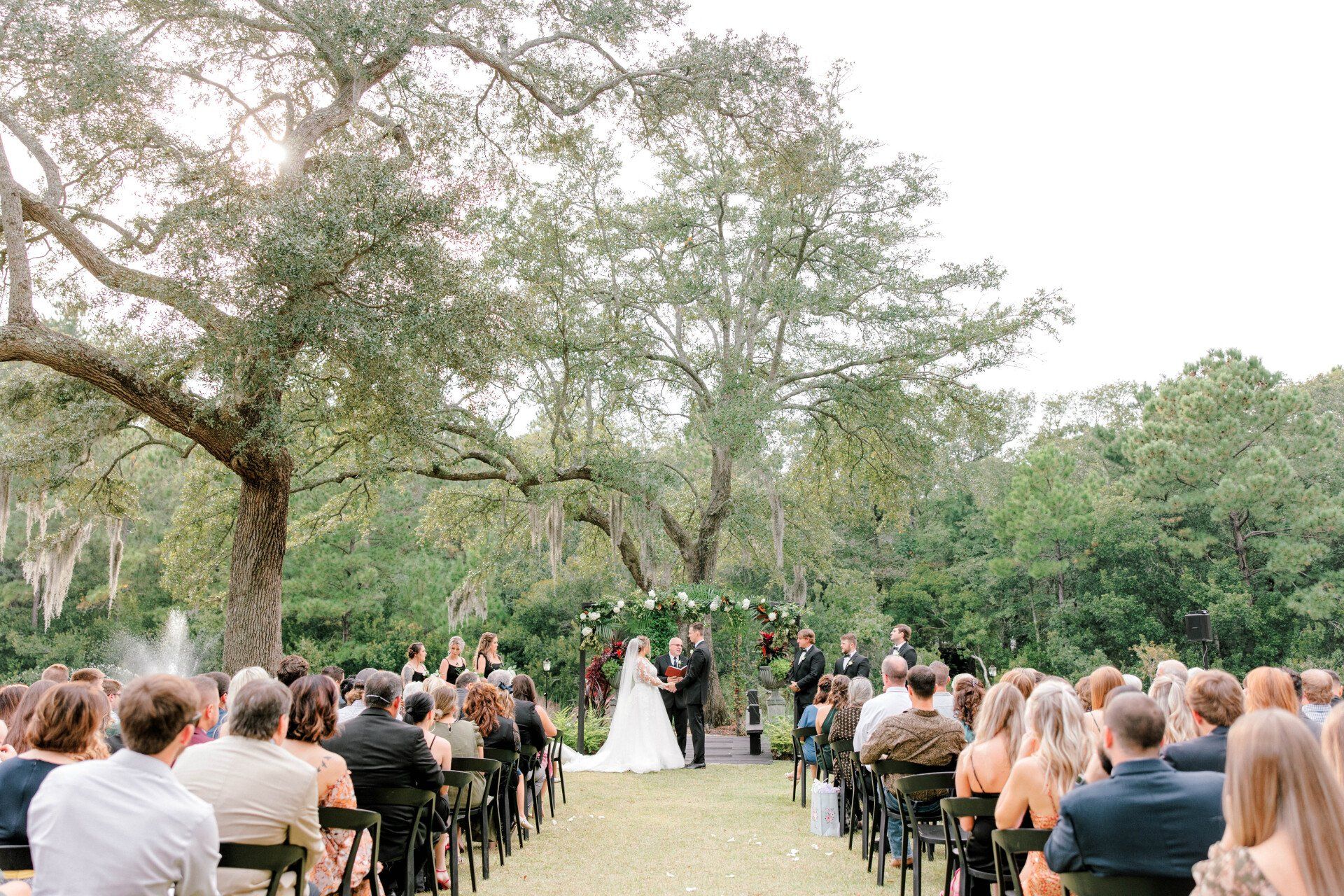 Outdoor wedding ceremony in a garden, with guests seated on both sides of an aisle under trees