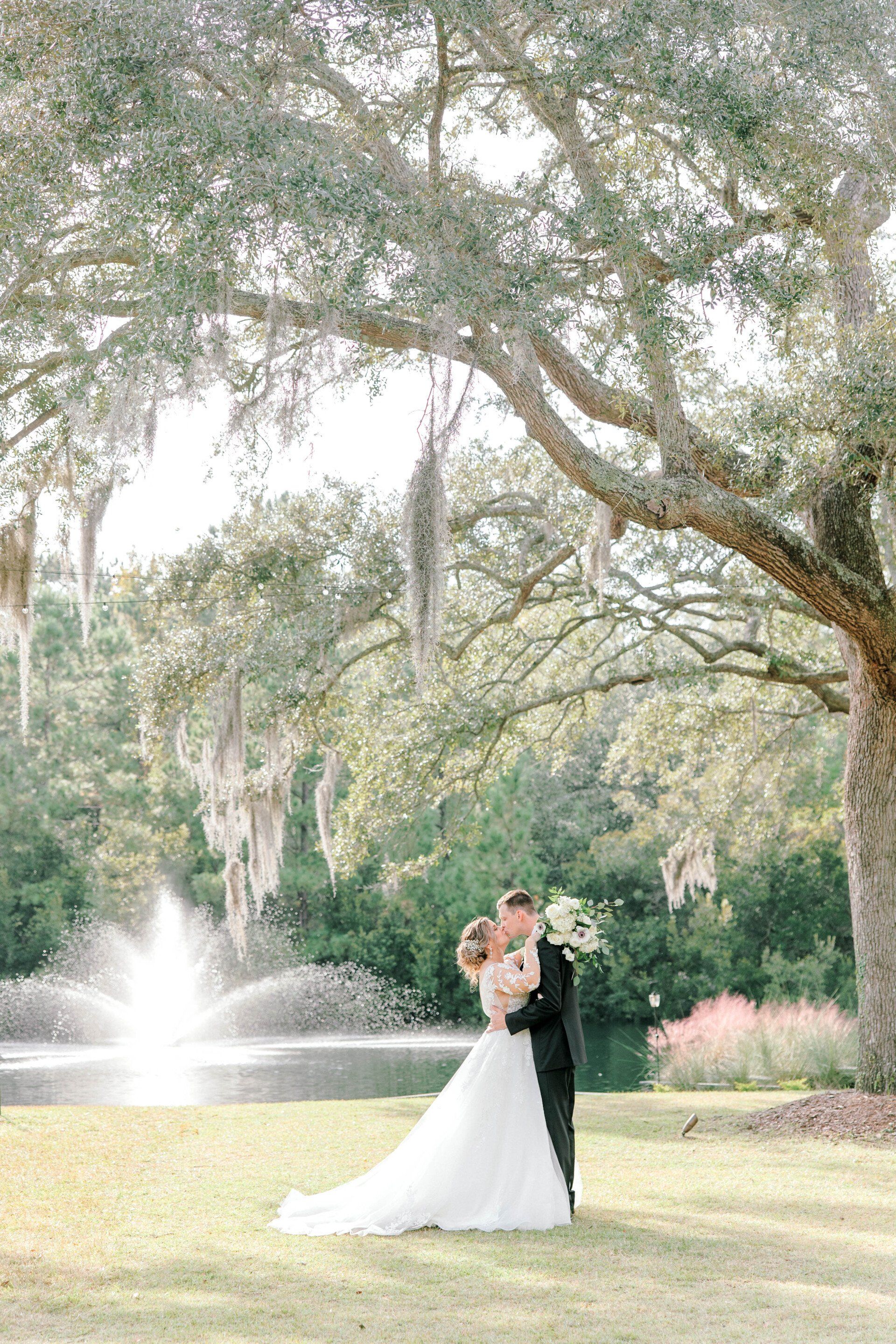 Bride and groom embracing under a large tree beside a fountain in a sunlit garden