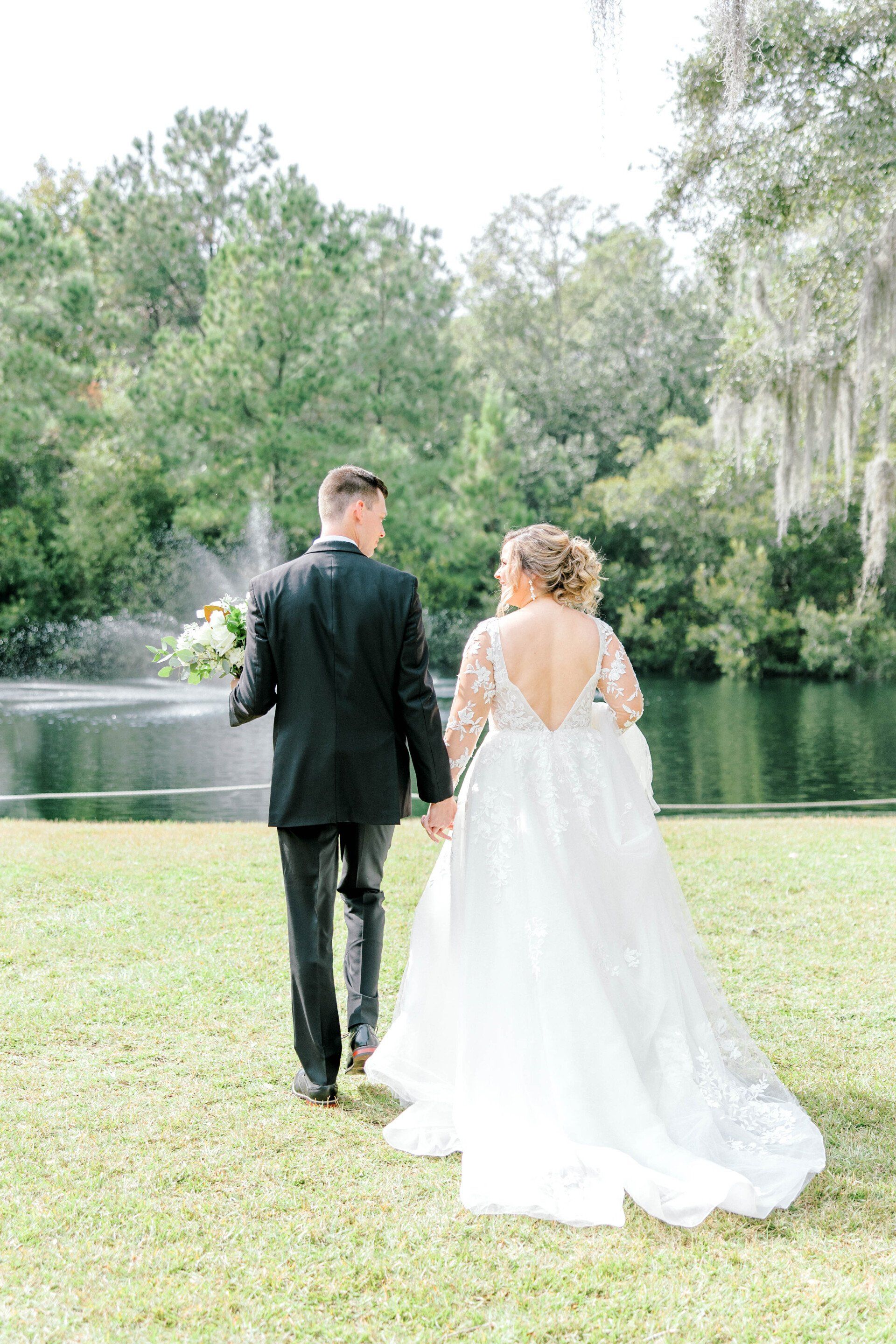 Couple in formal attire walking hand in hand by a lake in a green park