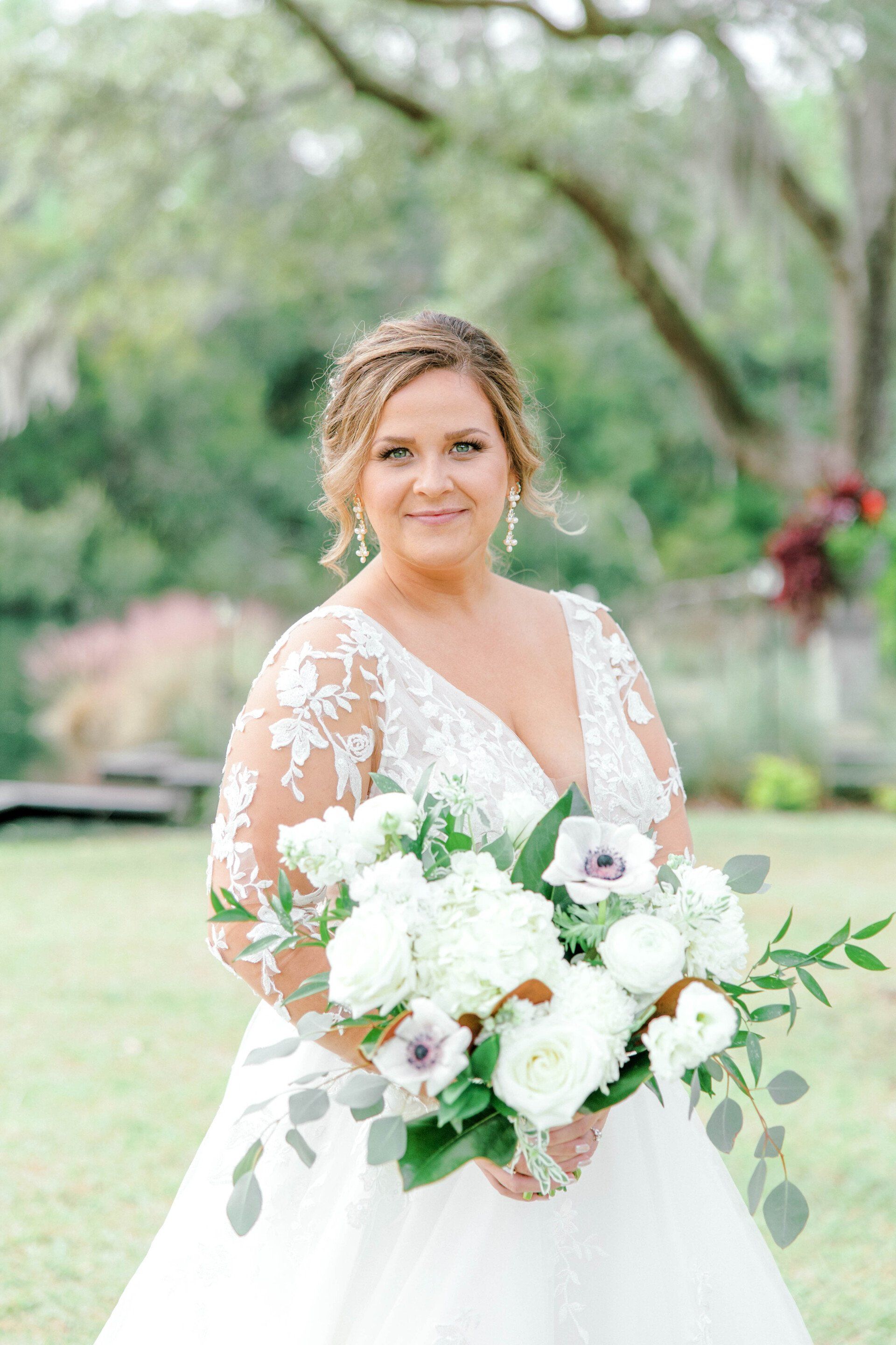 Bride in a white lace gown holding a white bouquet outdoors in a garden.
