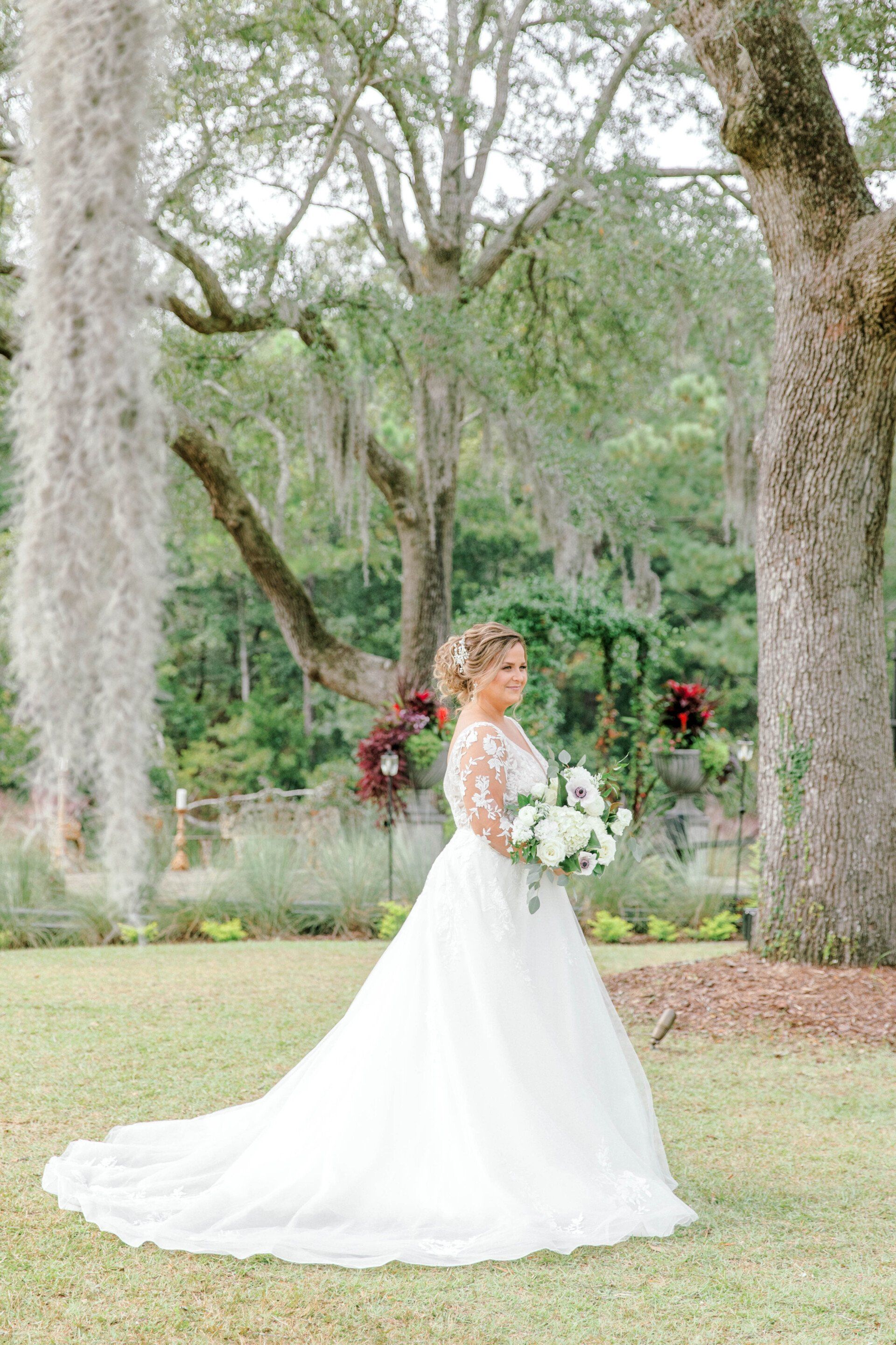 Bride in a white gown holding a bouquet in a garden under large oak trees