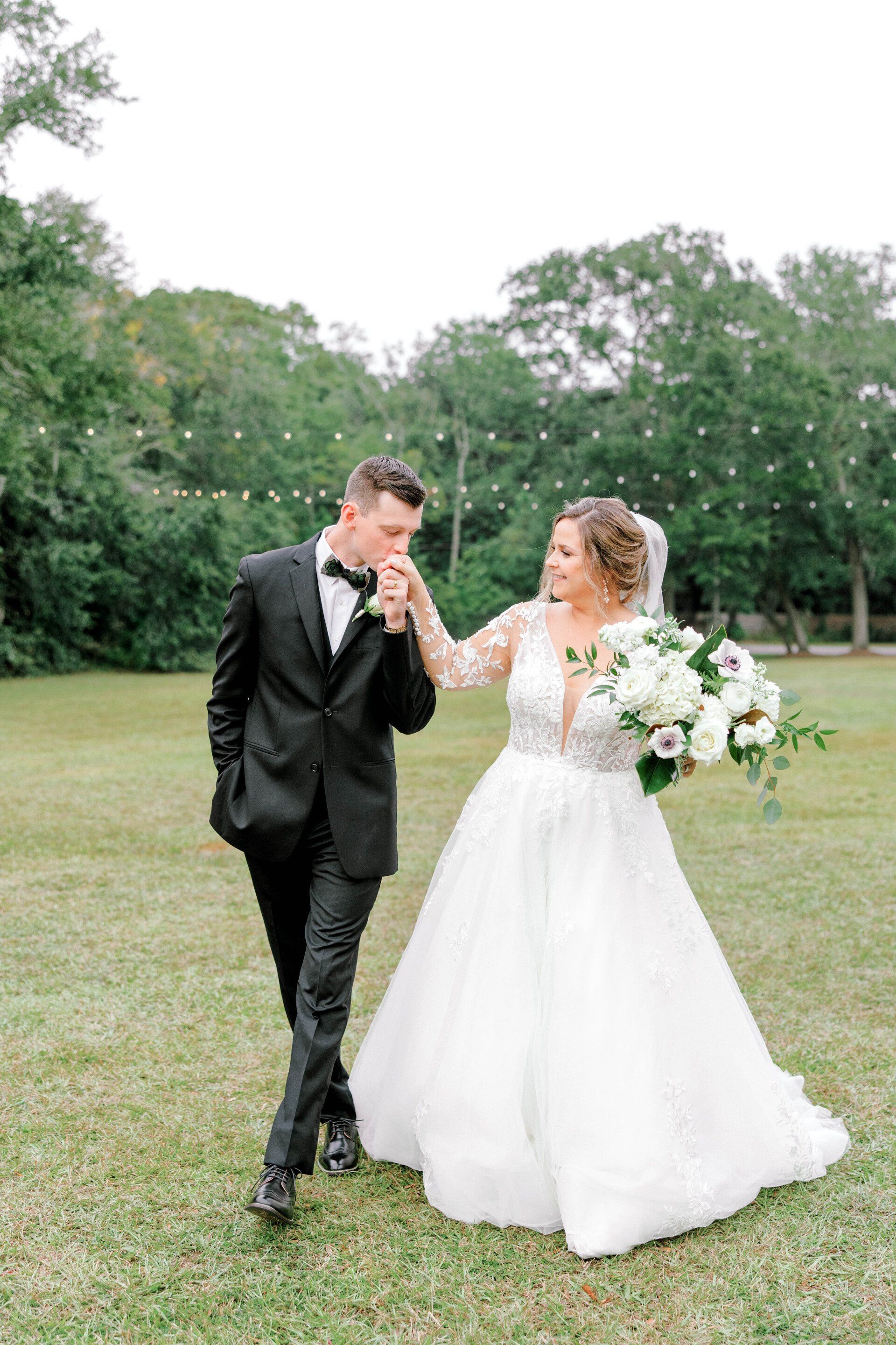Bride and groom kiss hands in a grassy park, holding a white floral bouquet.