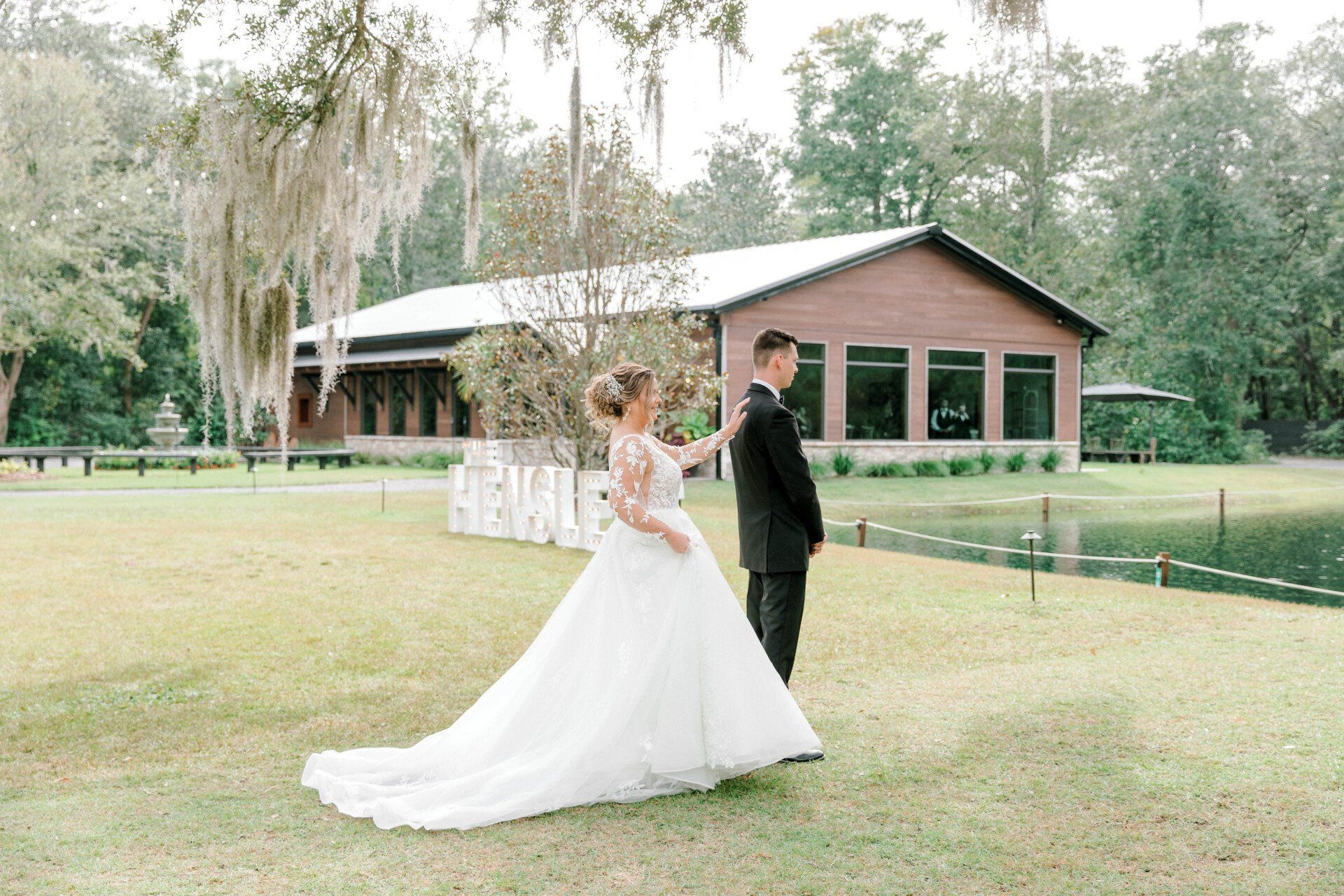 Bride in white gown and groom in black suit stand on a grassy lawn by a rustic building.