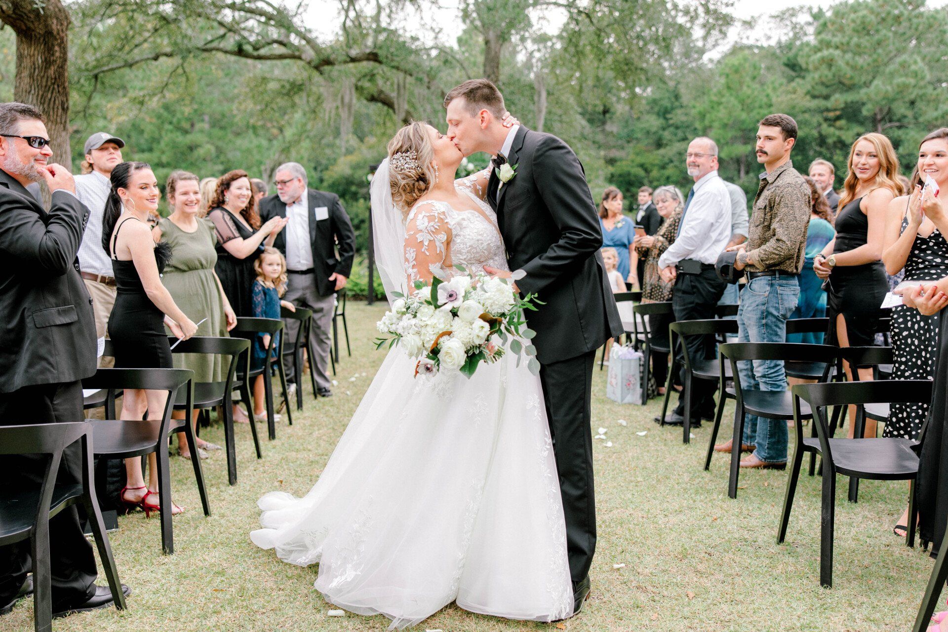 Bride and groom kiss in an outdoor wedding aisle, guests seated on both sides under trees.